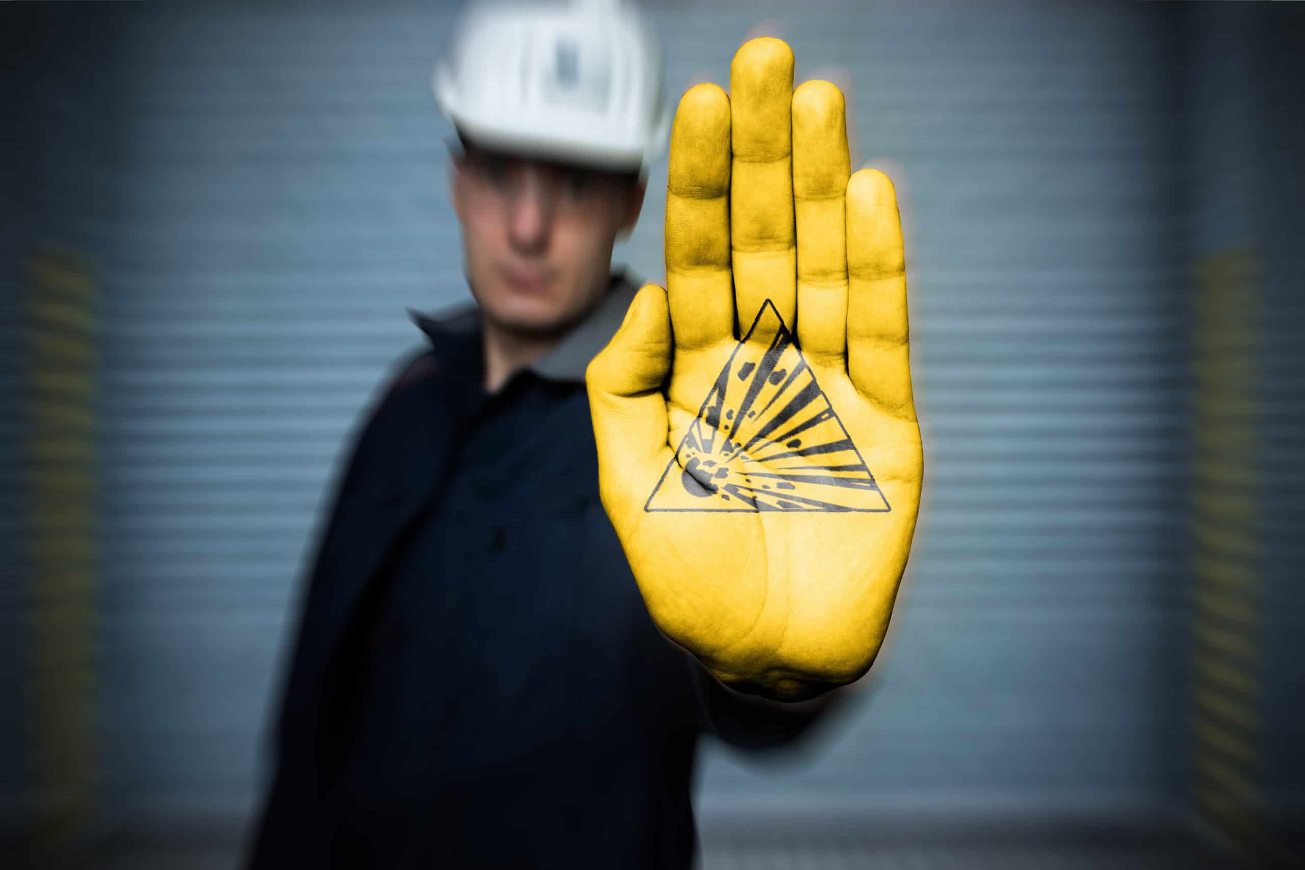 Mann mit Schutzhelm vor einem hellgrauen Hintergrund hebt seine Hand mit der Fläche nach vorne. Die Handfläche ist gelb angemalt und zeigt das Warnzeichen "Warnung vor explosionsgefährlichen Stoffen".