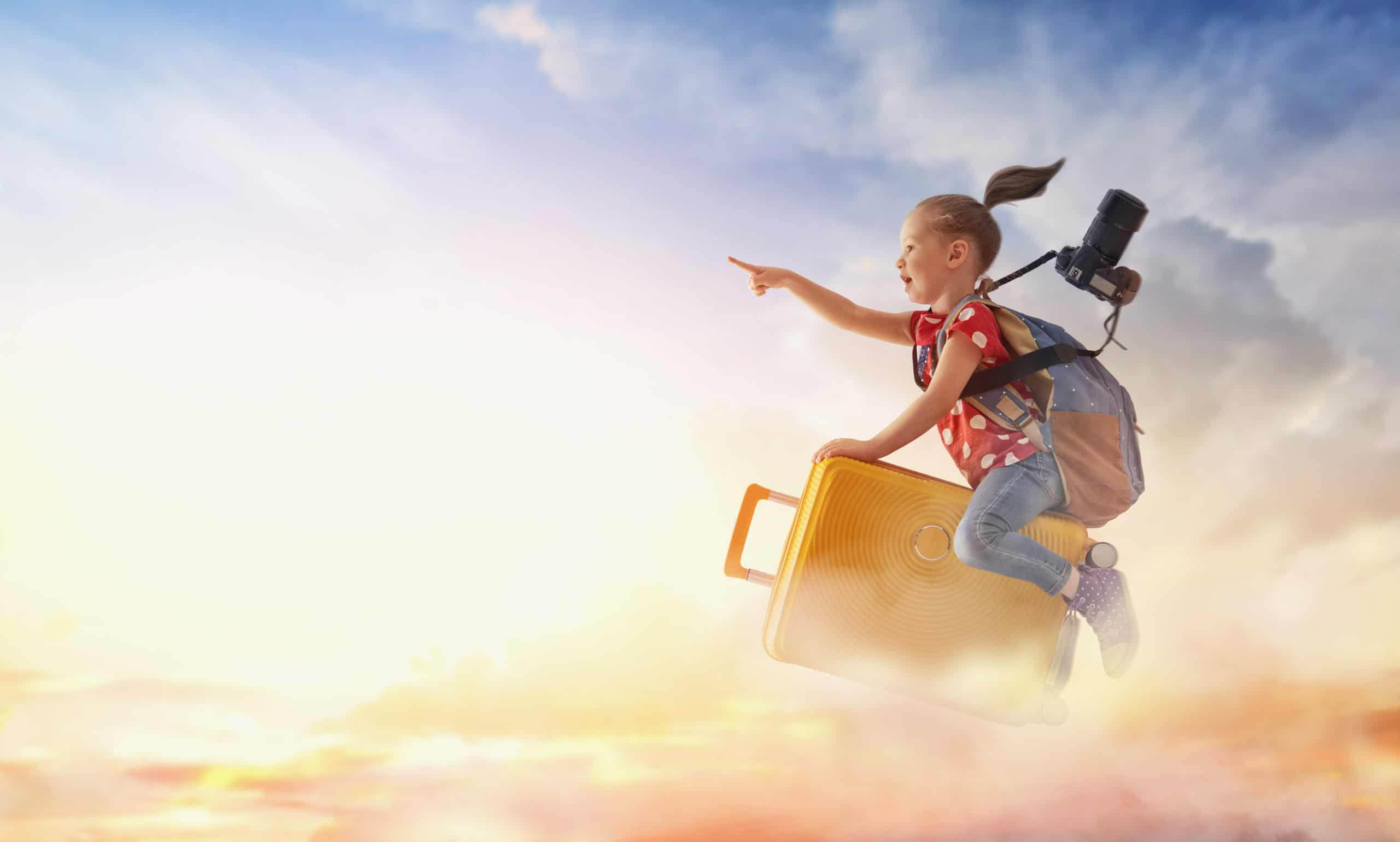 Kind mit Rucksack und großer Fotokamera sitzt auf einem Koffer und fliegt durch die Wolken in den Urlaub. Sie zeigt mit dem Finger nach vorne und ihr Zopf weht im Wind.