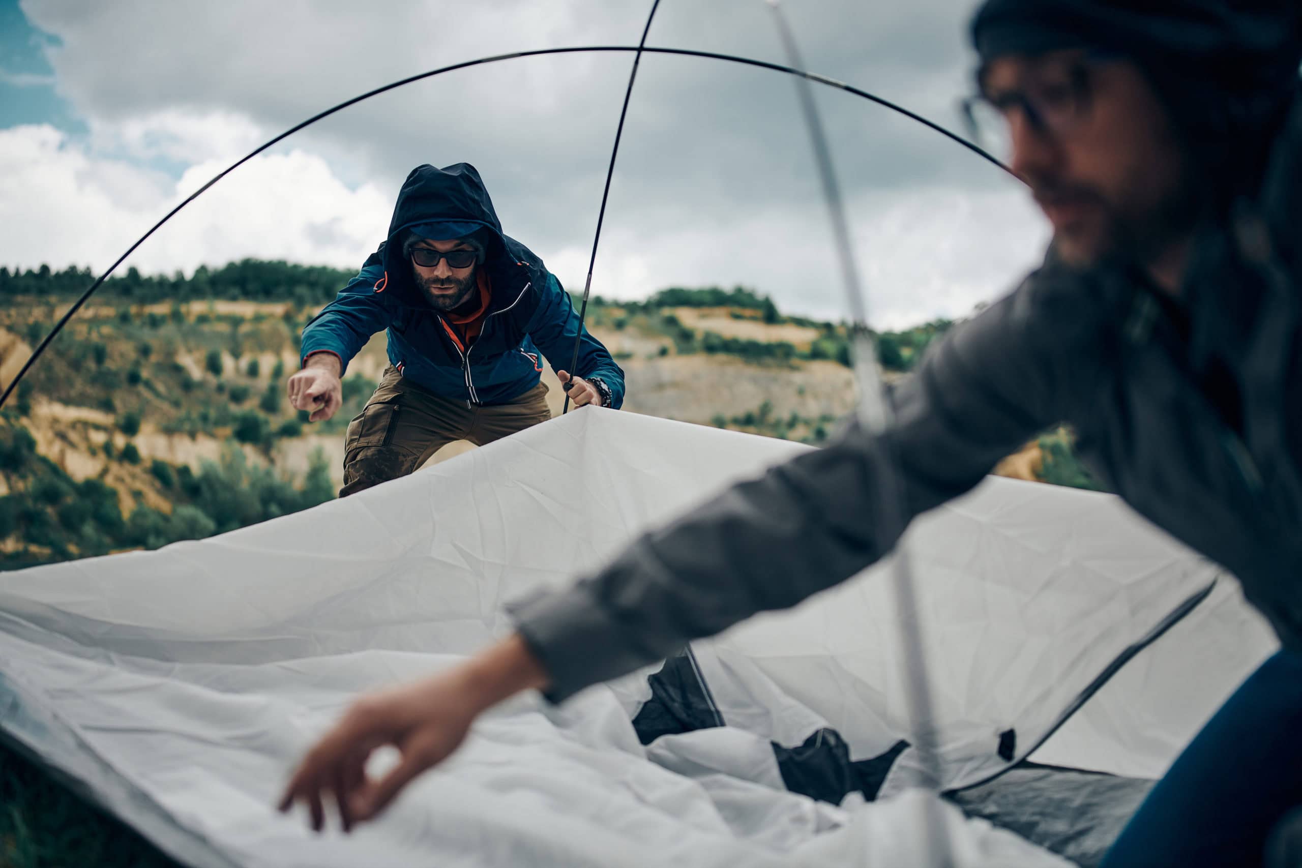 Zwei Männer mit Regenjacke bauen ein Zelt auf. Die Zeltstangen stehen bereits, das Zelt muss noch befestigt werden.