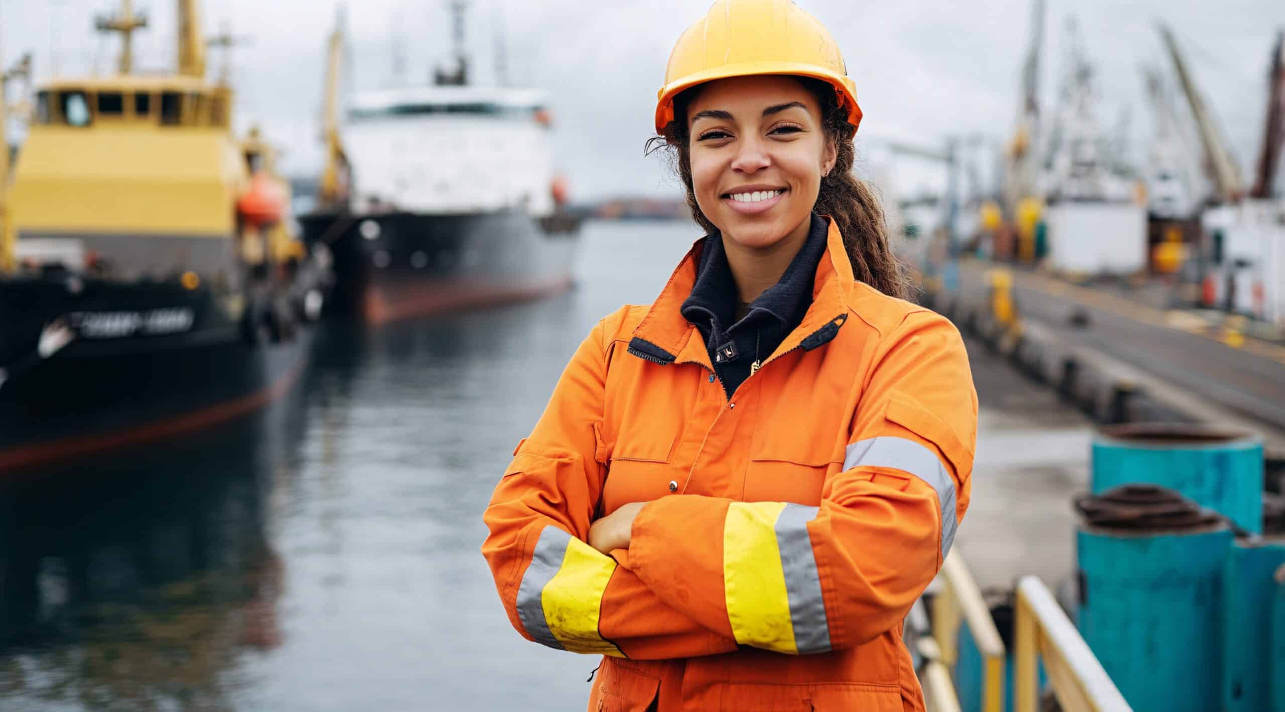 Frau mit Schutzhelm und orangener Sicherheitsjacke steht vor einem Hafen mit großen Schiffen